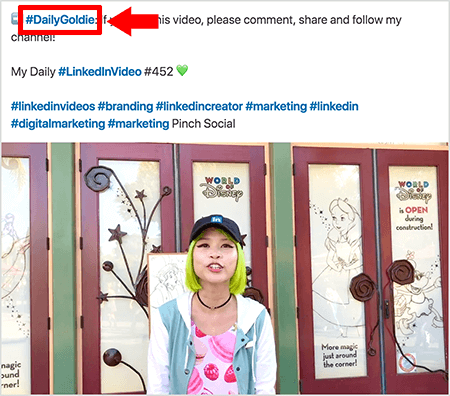 Dette er et skærmbillede, der illustrerer, hvordan Goldie Chan bruger hashtags i teksten i sine LinkedIn-videoindlæg. Røde billedtekster peger på hashtagget #DailyGoldie i teksten, hvilket er unikt for hendes videoindlæg og hjælper hende med at spore aktier. Indlægget indeholder også andre relevante hashtags, der hjælper folk med at finde hendes video, herunder #LinkedInVideo. I videobilledet står Goldie foran nogle døre ved en World of Disney-skærm. Hun er en asiatisk kvinde med grønt hår. Hun har en sort LinkedIn-kasket, en sort chokerhalskæde, en lyserød macaron-print-skjorte og en blå og hvid jakke.